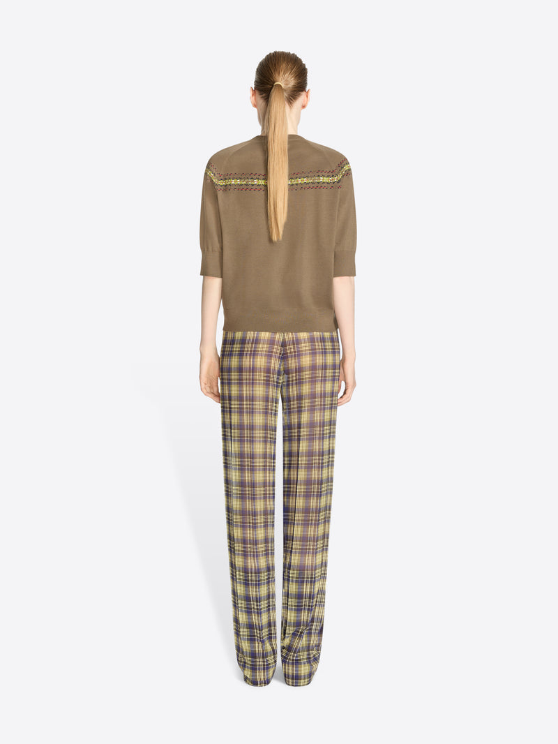 Back view of a model standing against a light gray background wearing a brown knit sweater with a colorful geometric pattern stripe across the chest and shoulders, olive green and yellow plaid trousers, and dark brown shoes.