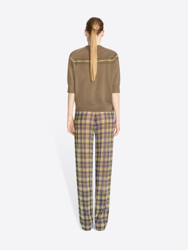 Back view of a model standing against a light gray background wearing a brown knit sweater with a colorful geometric pattern stripe across the chest and shoulders, olive green and yellow plaid trousers, and dark brown shoes.