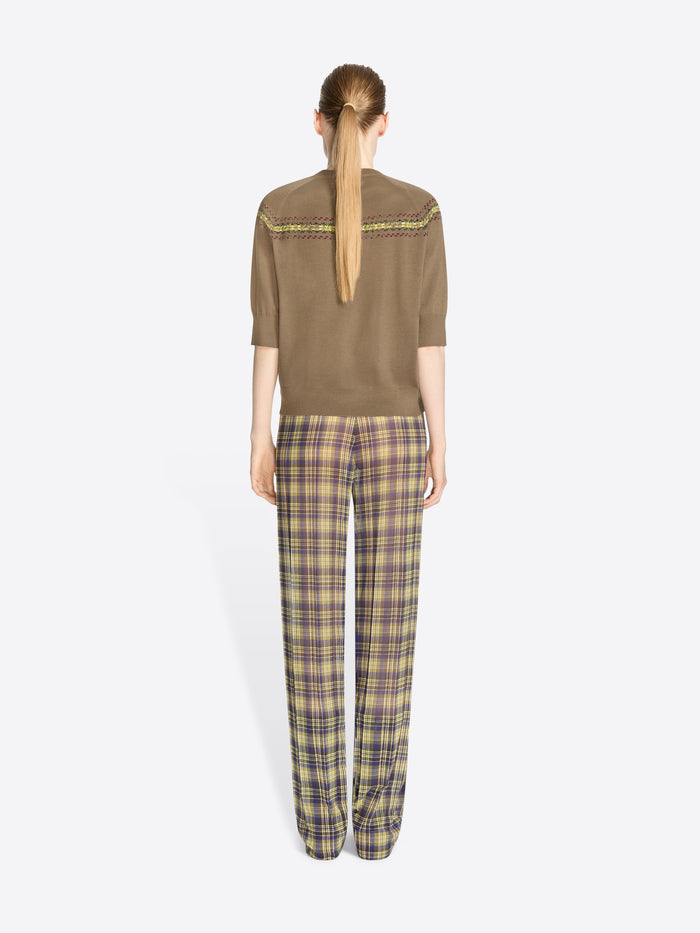 Back view of a model standing against a light gray background wearing a brown knit sweater with a colorful geometric pattern stripe across the chest and shoulders, olive green and yellow plaid trousers, and dark brown shoes.