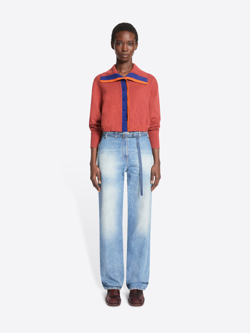 A model stands against a light gray background wearing a cropped, long-sleeve knit cardigan with a purple center placket, and a wide collar with purple and orange accents, paired with light-wash wide-leg jeans and dark brown shoes.