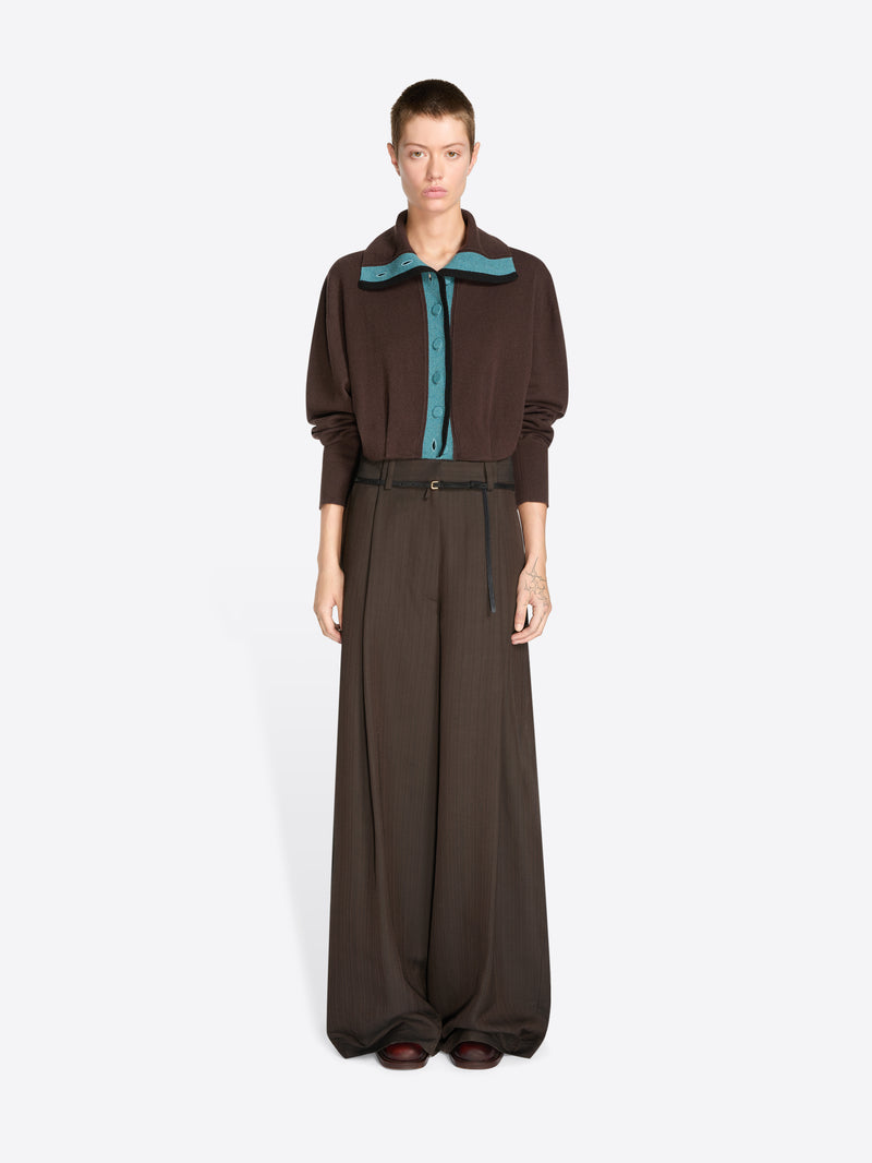 A model stands against a light gray background wearing a dark brown long-sleeve collared cardigan with a teal inner placket and matching wide-leg trousers with a thin waist belt.