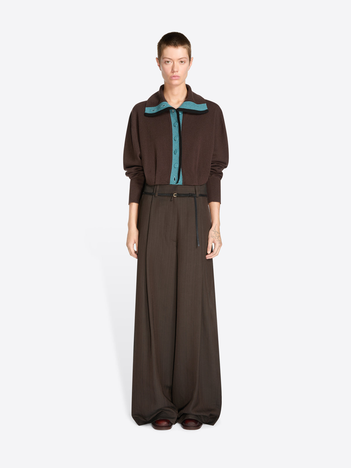 A model stands against a light gray background wearing a dark brown long-sleeve collared cardigan with a teal inner placket and matching wide-leg trousers with a thin waist belt.