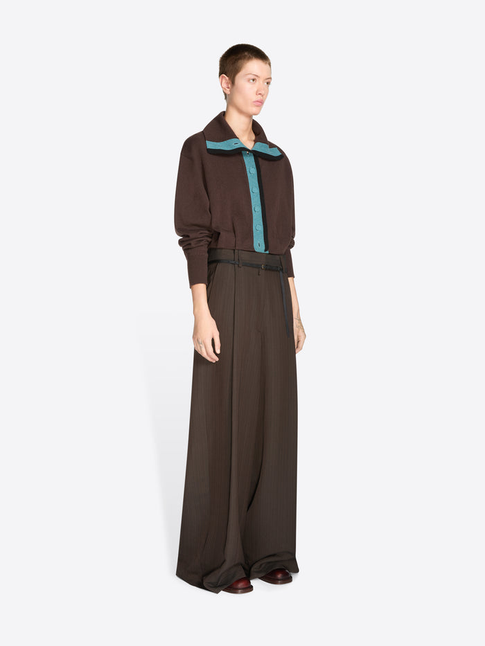 A model stands against a light gray background wearing a dark brown long-sleeve collared cardigan with a teal inner placket and matching wide-leg trousers with a thin waist belt.