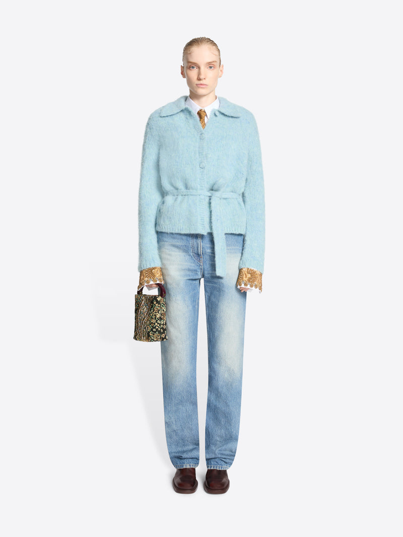 A model stands against a light gray background wearing a light blue belted cardigan over a shirt with gold-patterned cuffs, light-wash blue jeans, dark brown shoes, and holding a small patterned handbag.