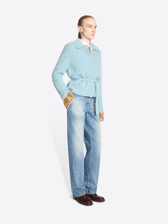 A model stands against a light gray background wearing a light blue belted cardigan over a shirt with gold-patterned cuffs, light-wash blue jeans and dark brown shoes.
