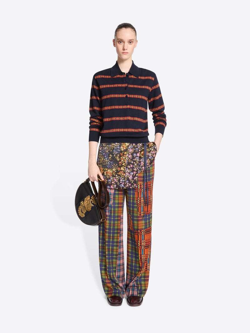 A model stands against a light gray background wearing a navy and orange striped knit polo sweater, wide-leg patchwork plaid pants, skirt, and holding a black round handbag with gold detail.