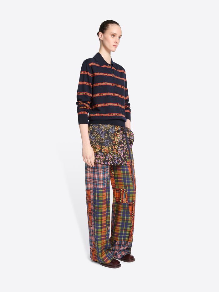 A model stands against a light gray background wearing a navy and orange striped knit polo sweater, wide-leg patchwork plaid pants and skirt.