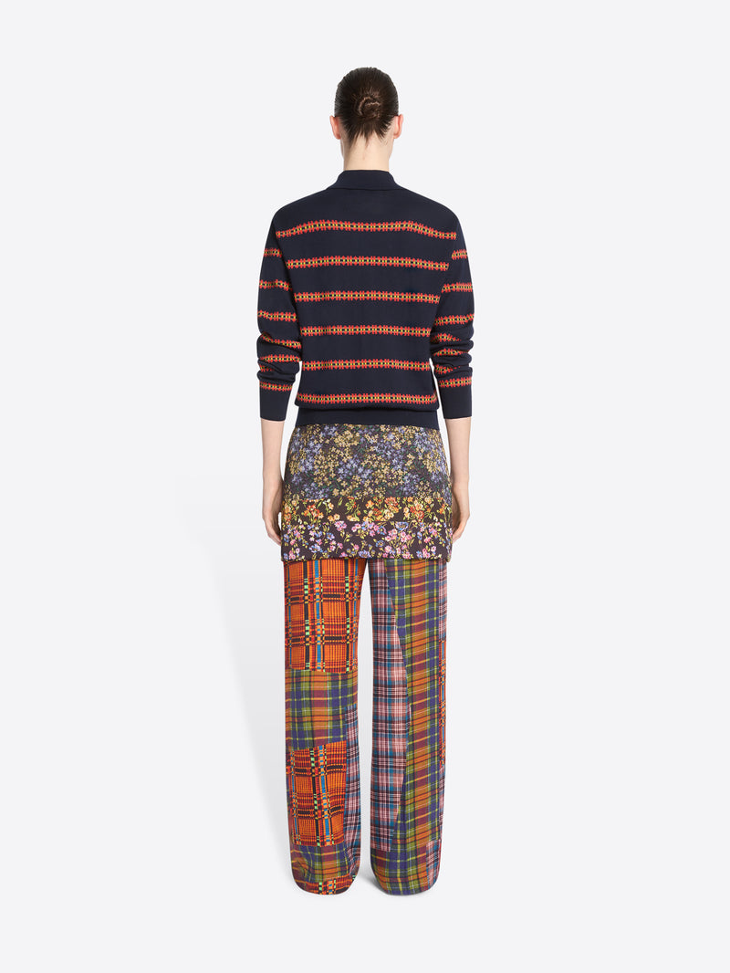 Back view of a model standing against a light gray background wearing a navy and orange striped knit polo sweater, wide-leg patchwork plaid trousers and skirt.