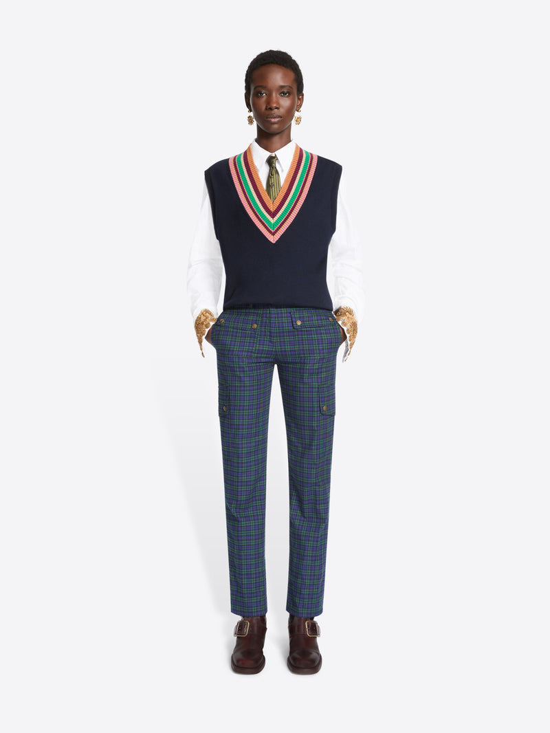 A model stands against a light gray background wearing a navy sweater vest with a colorful striped V-neck trim over a white collared shirt and tie, paired with navy plaid trousers and dark brown shoes.
