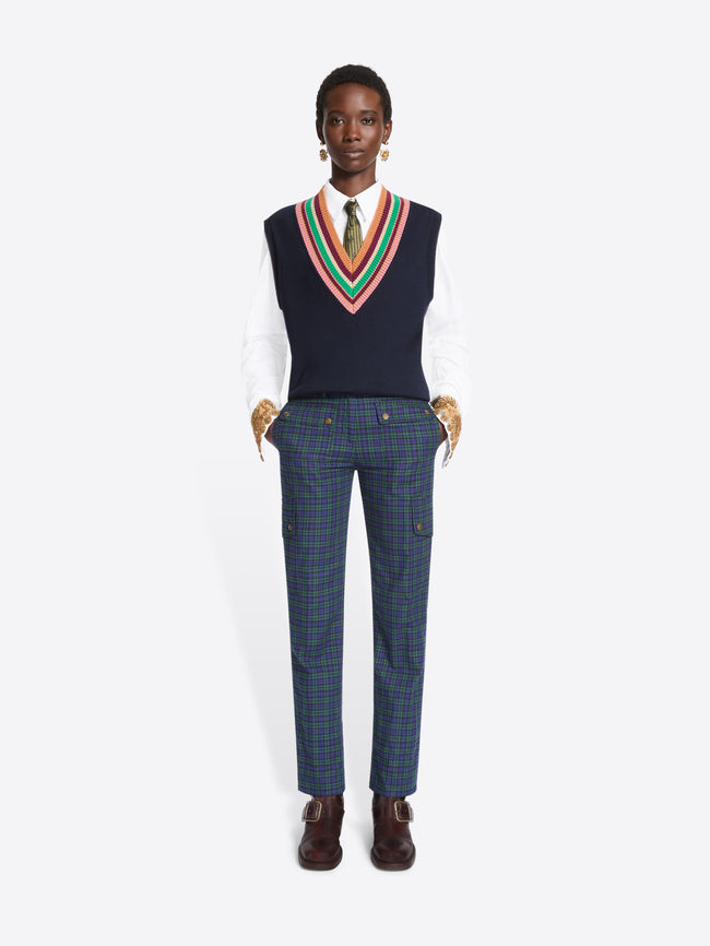 A model stands against a light gray background wearing a navy sweater vest with a colorful striped V-neck trim over a white collared shirt and tie, paired with navy plaid trousers and dark brown shoes.