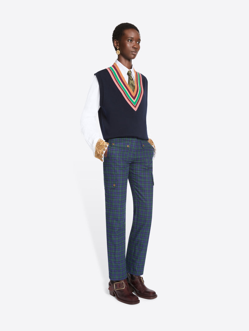A model stands against a light gray background wearing a navy sweater vest with a colorful striped V-neck trim over a white collared shirt and tie, paired with navy plaid trousers and dark brown shoes.