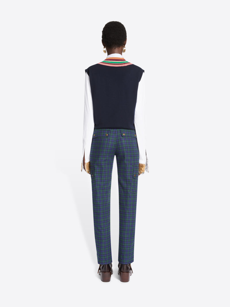 Back view of a model standing against a light gray background wearing a navy sweater vest with a colorful striped V-neck trim over a white collared shirt and tie, paired with navy plaid trousers and dark brown shoes.