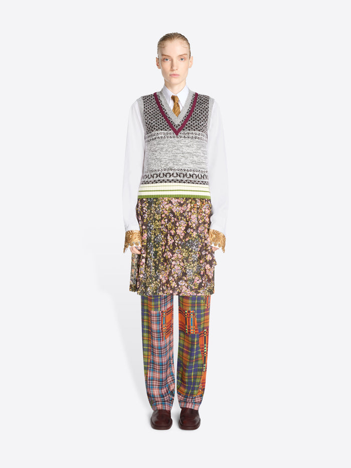A model stands against a light gray background wearing a layered outfit: a white long-sleeve shirt, a gray patterned V-neck sweater vest, a floral printed skirt, and plaid trousers in multiple colors.
