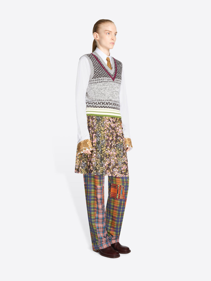 A model stands against a light gray background wearing a layered outfit: a white long-sleeve shirt, a gray patterned V-neck sweater vest, a floral printed skirt, and plaid trousers in multiple colors.
