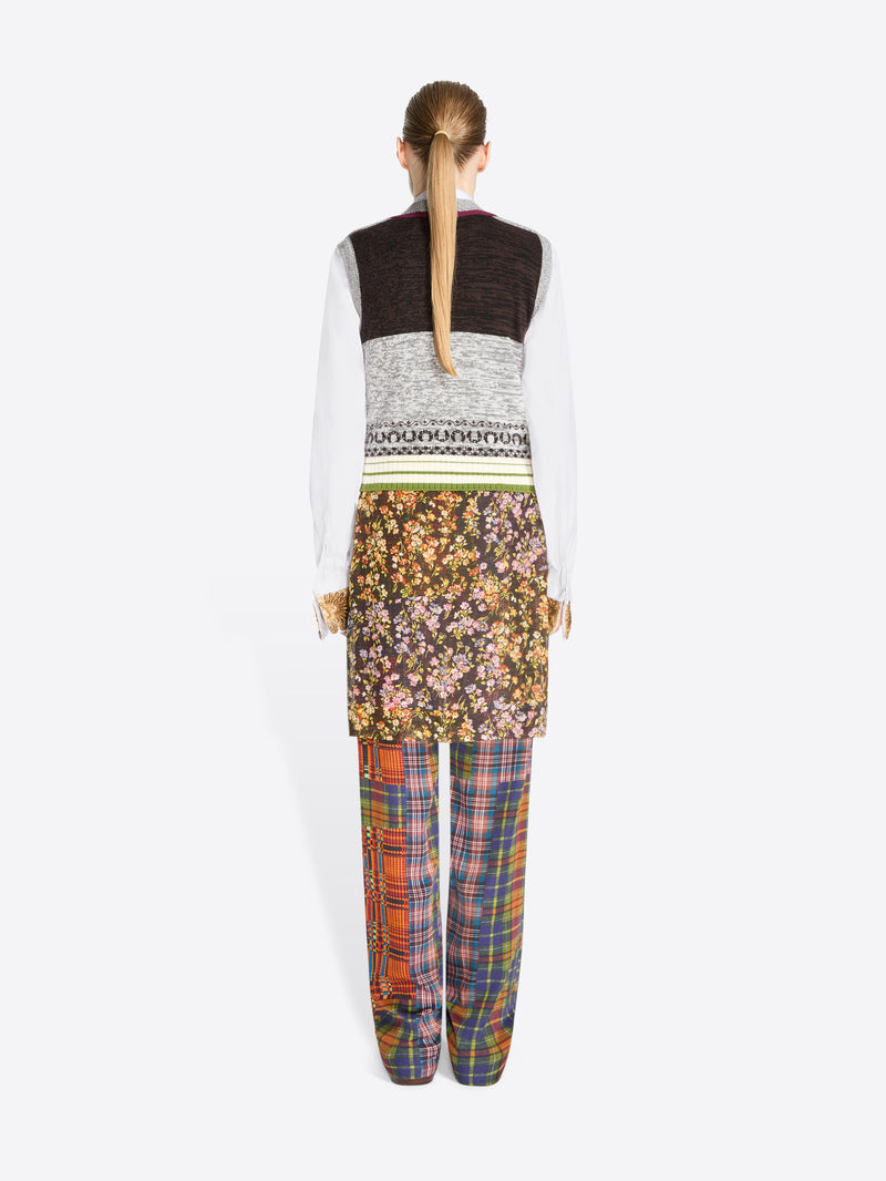 Back view of a model standing against a light gray background wearing a layered outfit: a white long-sleeve shirt, a gray patterned V-neck sweater vest, a floral printed skirt, and plaid trousers in multiple colors.