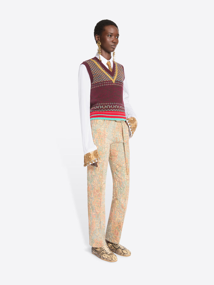A model stands against a light gray background wearing a white long-sleeve shirt, a burgundy and gold patterned knit sweater vest, a white tie, beige patterned trousers, and snakeskin shoes.