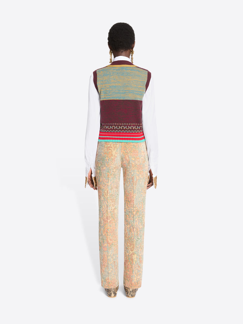 Back view of a model against a light gray background wearing a white long-sleeve shirt, a burgundy and gold patterned knit sweater vest, a white tie, beige patterned trousers, and snakeskin shoes.