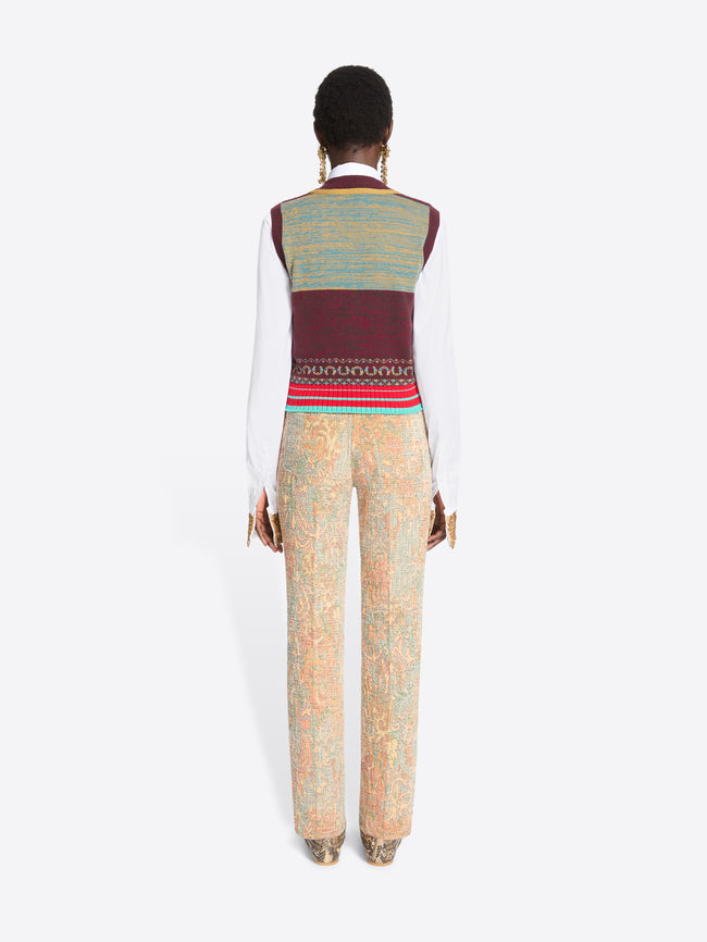 Back view of a model against a light gray background wearing a white long-sleeve shirt, a burgundy and gold patterned knit sweater vest, a white tie, beige patterned trousers, and snakeskin shoes.