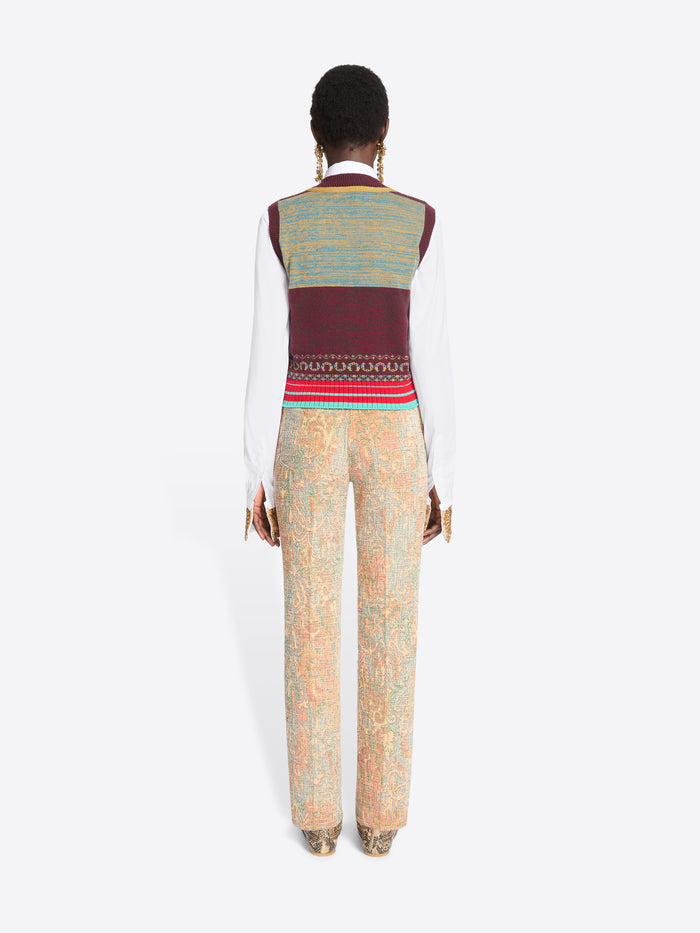 Back view of a model against a light gray background wearing a white long-sleeve shirt, a burgundy and gold patterned knit sweater vest, a white tie, beige patterned trousers, and snakeskin shoes.