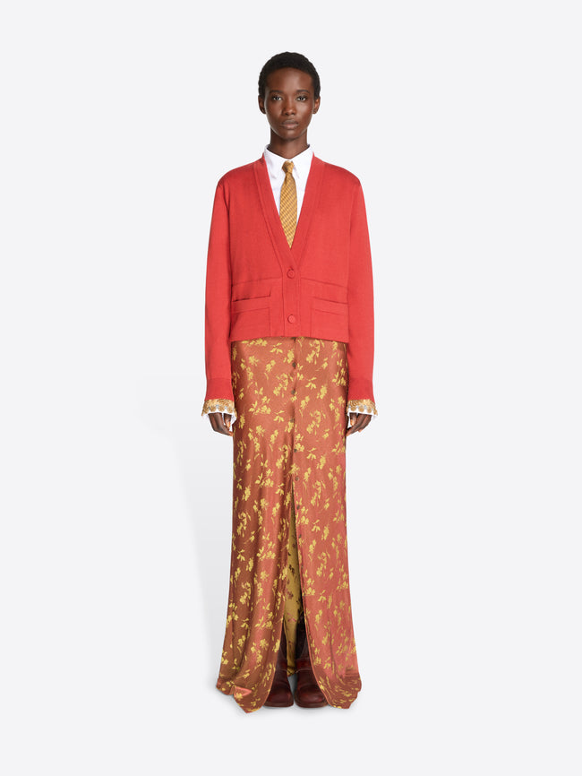 A model stands against a light gray background wearing a bright red cropped cardigan over a white collared shirt and a gold patterned tie, paired with rust-orange patterned skirt and dark brown shoes.