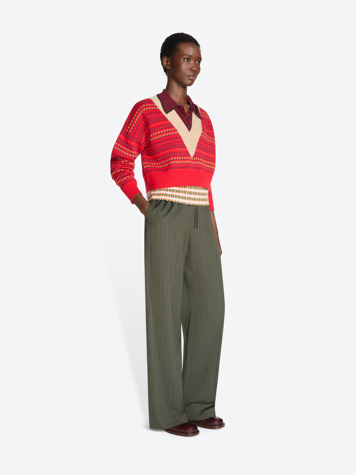 A model stands against a light gray background wearing a red and beige patterned knit sweater, paired with olive green wide-leg pants.