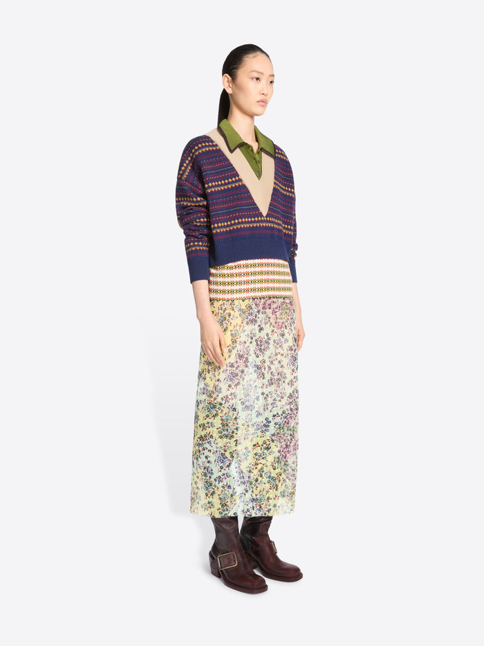 A model stands against a light gray background wearing a patterned knit sweater with an olive collar, a long floral print skirt and dark brown ankle boots.