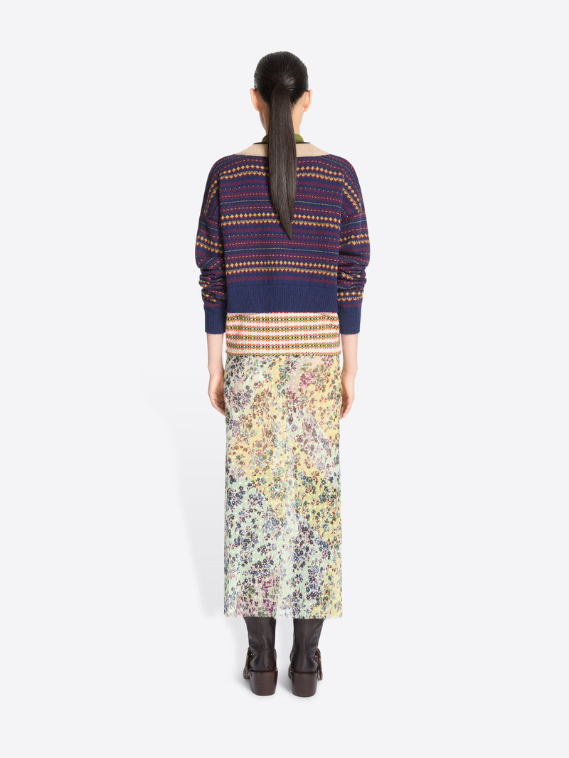 Back view of a model standing against a light gray background wearing a patterned knit sweater with an olive collar, a long floral print skirt, dark brown ankle boots, and holding a colorful oval handbag.