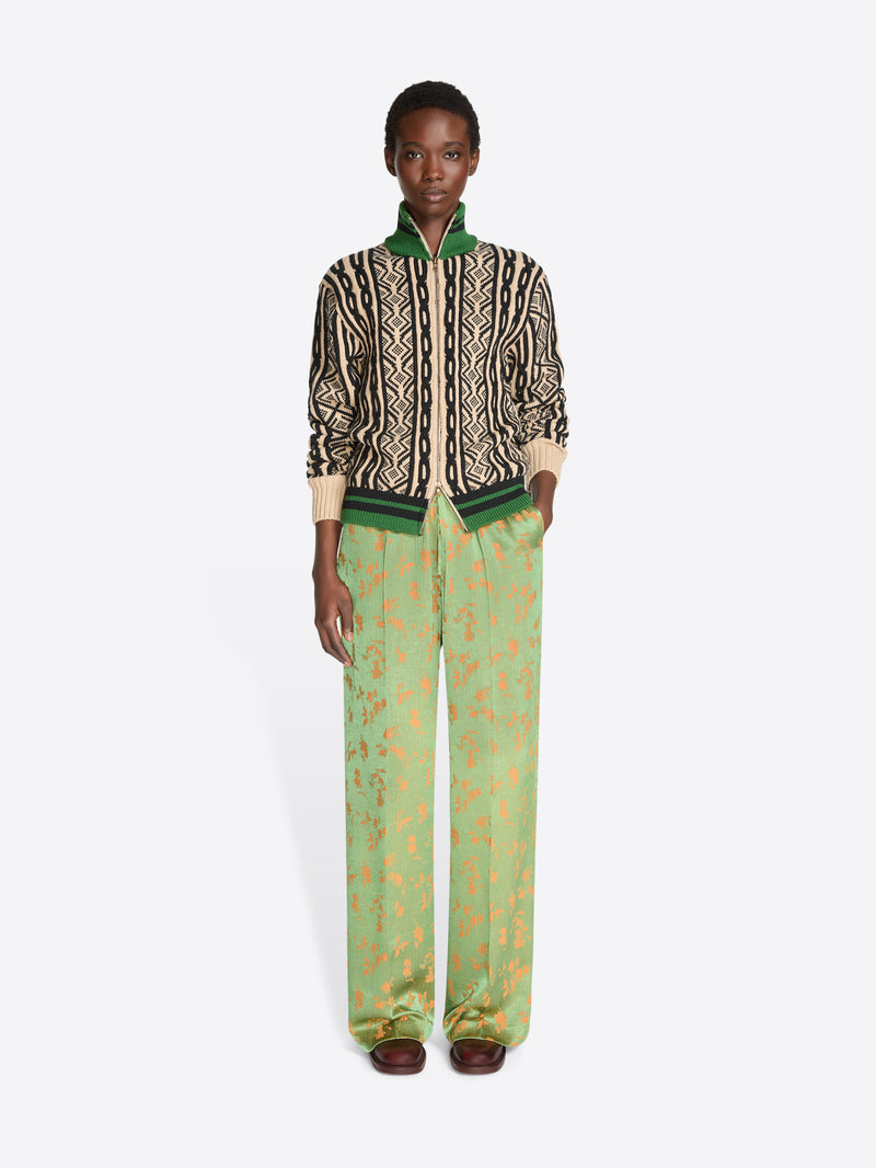 A model stands against a light gray background wearing a cropped knit jacket with a geometric pattern, paired with wide-leg green trousers featuring a gold abstract print.