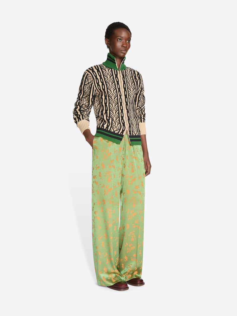 A model stands against a light gray background wearing a cropped knit jacket with a geometric pattern, paired with wide-leg green trousers featuring a gold abstract print.