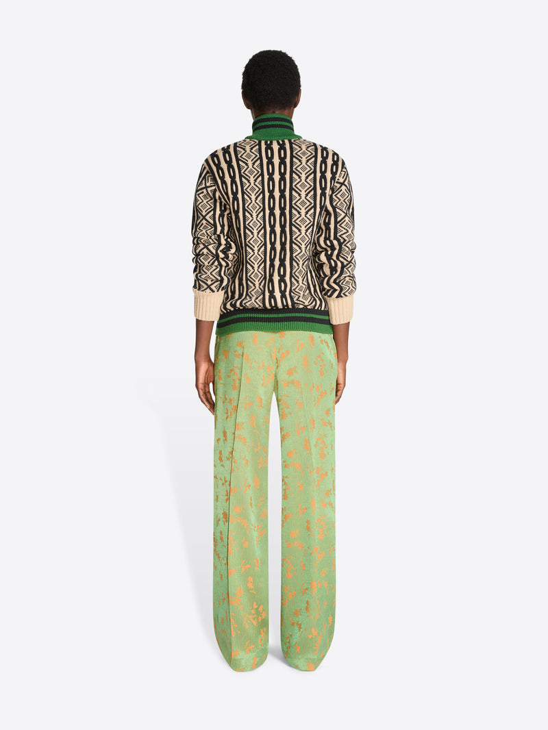 Back view of a model standing against a light gray background wearing a cropped knit jacket with a geometric pattern, paired with wide-leg green trousers featuring a gold abstract print.