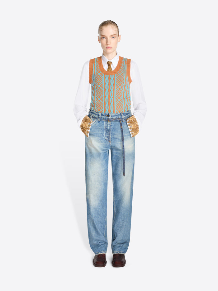 A model stands against a light gray background wearing a white long-sleeve shirt, a patterned orange and blue knit vest, light blue jeans with a belt at the waist, and dark brown shoes.
