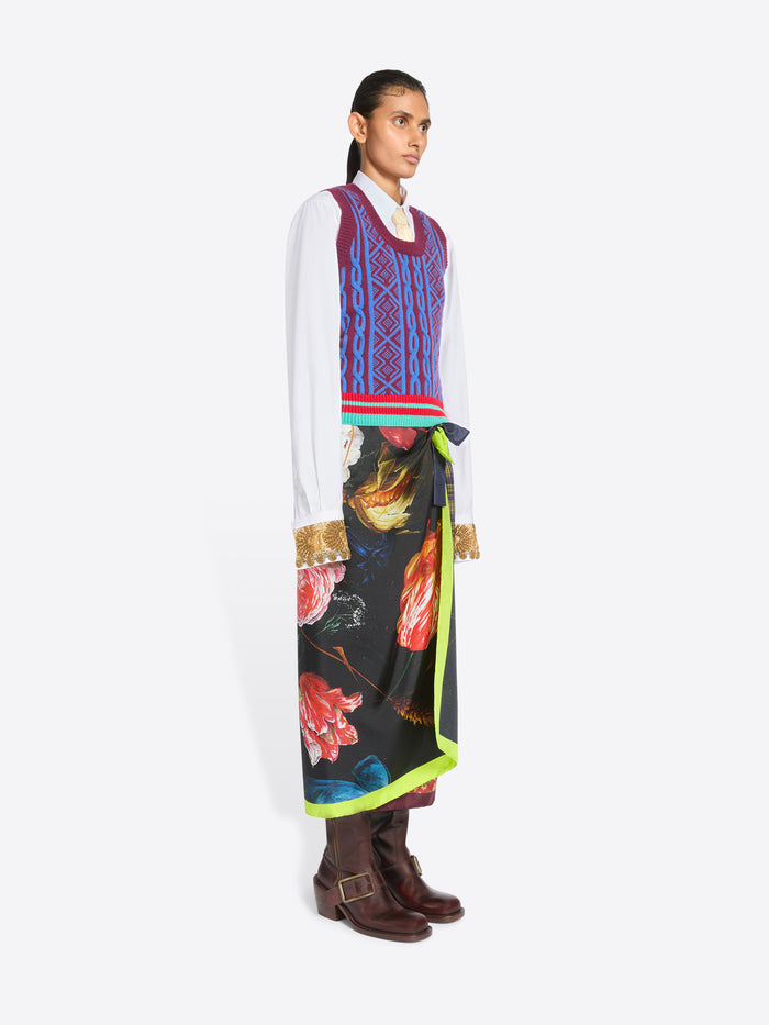 A model stands against a light gray background wearing a white long-sleeve shirt, a colorful patterned knit sweater vest, a long multicolor skirt with a bright neon green side panel, and dark brown leather boots, holding a black bag with gold detailing.