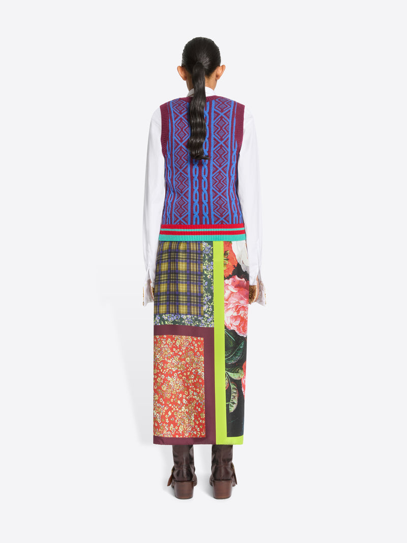 Back view of a model standing against a light gray background wearing a white long-sleeve shirt, a colorful patterned knit sweater vest, a long multicolor skirt with a bright neon green side panel, and dark brown leather boots, holding a black bag with gold detailing.