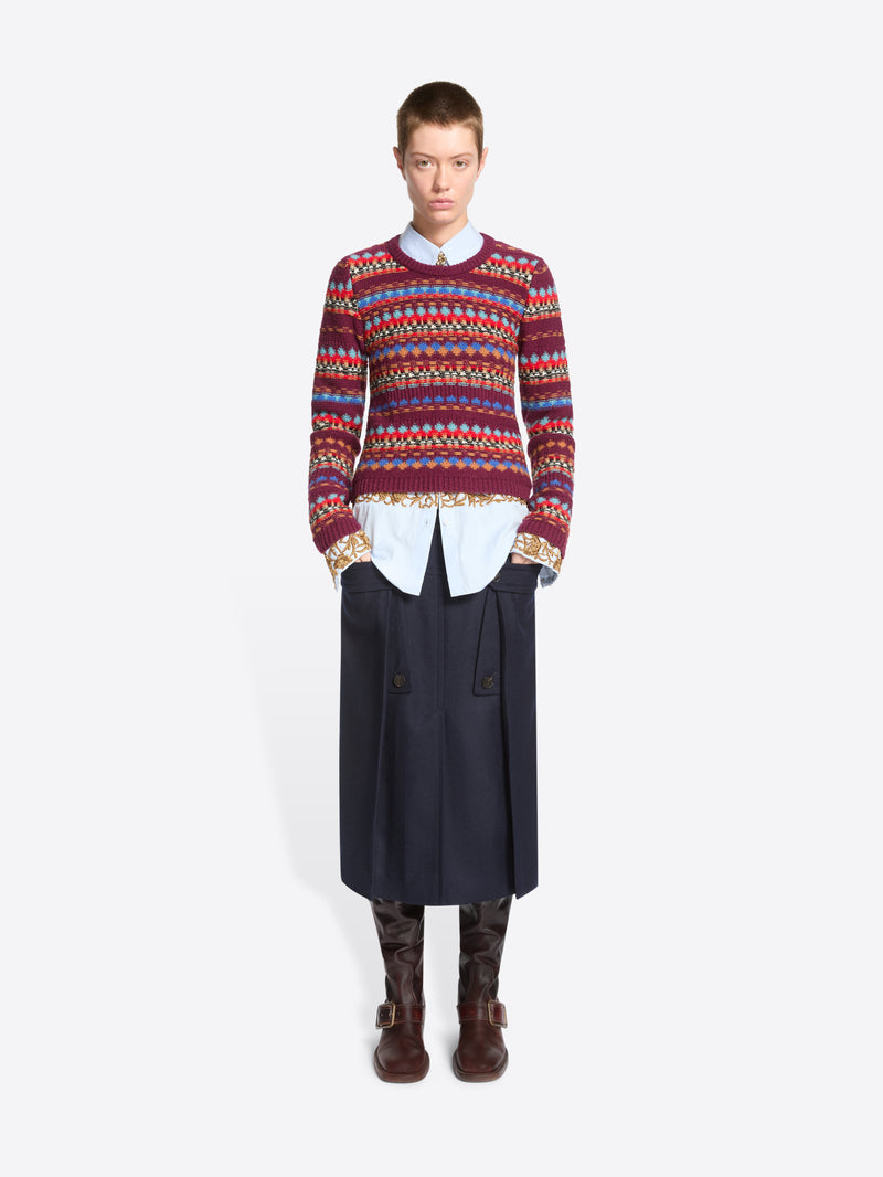 A model stands against a light gray background wearing a long-sleeve crewneck sweater with a multicolor geometric pattern, layered over a white collared shirt and dark navy skirt.