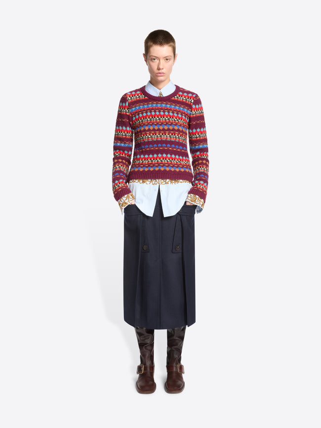 A model stands against a light gray background wearing a long-sleeve crewneck sweater with a multicolor geometric pattern, layered over a white collared shirt and dark navy skirt.
