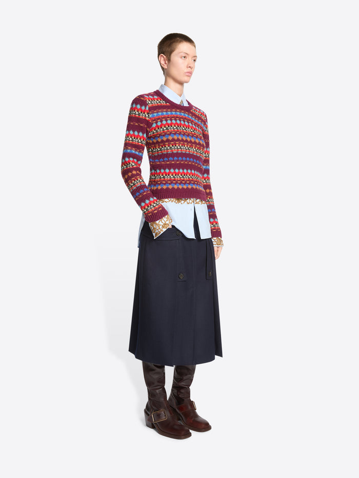 A model stands against a light gray background wearing a long-sleeve crewneck sweater with a multicolor geometric pattern, layered over a white collared shirt and dark navy skirt.