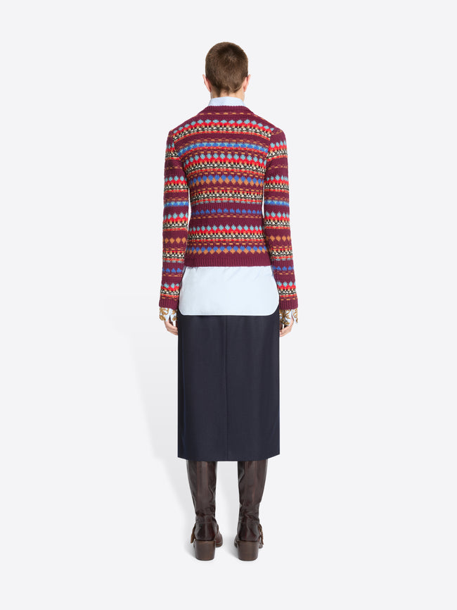 Back view of a model standing against a light gray background wearing a long-sleeve crewneck sweater with a multicolor geometric pattern, layered over a white collared shirt and dark navy skirt.