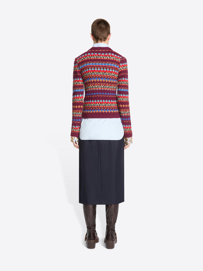 Back view of a model standing against a light gray background wearing a long-sleeve crewneck sweater with a multicolor geometric pattern, layered over a white collared shirt and dark navy skirt.