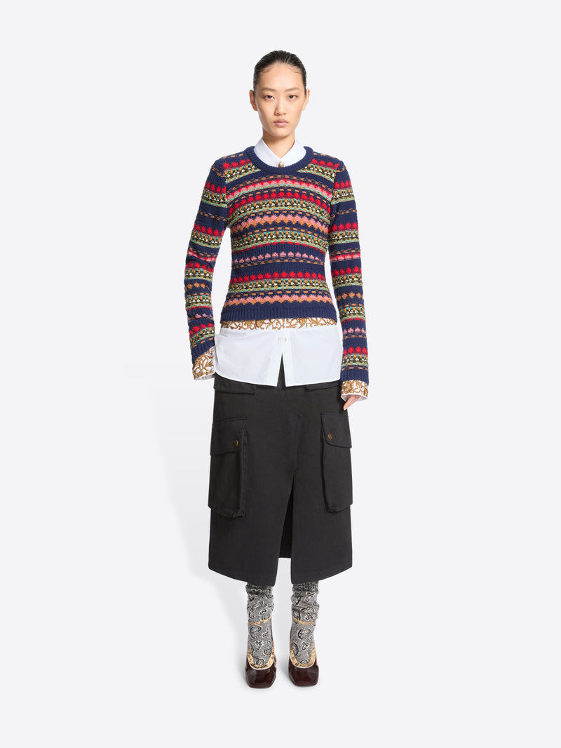 A model stands against a light gray background wearing a long-sleeve crewneck sweater with multicolor geometric stripes, a white collared shirt underneath, a black midi skirt with cargo pockets, patterned socks, and dark brown heels.
