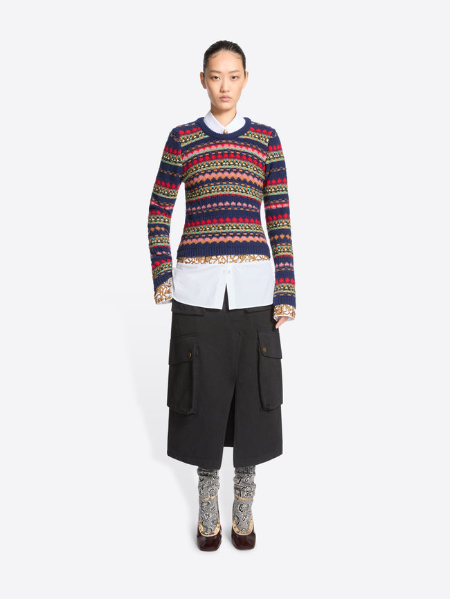 A model stands against a light gray background wearing a long-sleeve crewneck sweater with multicolor geometric stripes, a white collared shirt underneath, a black midi skirt with cargo pockets, patterned socks, and dark brown heels.