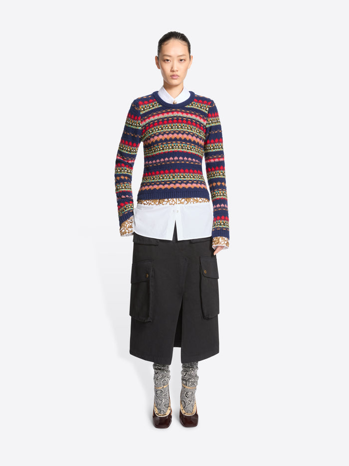 A model stands against a light gray background wearing a long-sleeve crewneck sweater with multicolor geometric stripes, a white collared shirt underneath, a black midi skirt with cargo pockets, patterned socks, and dark brown heels.