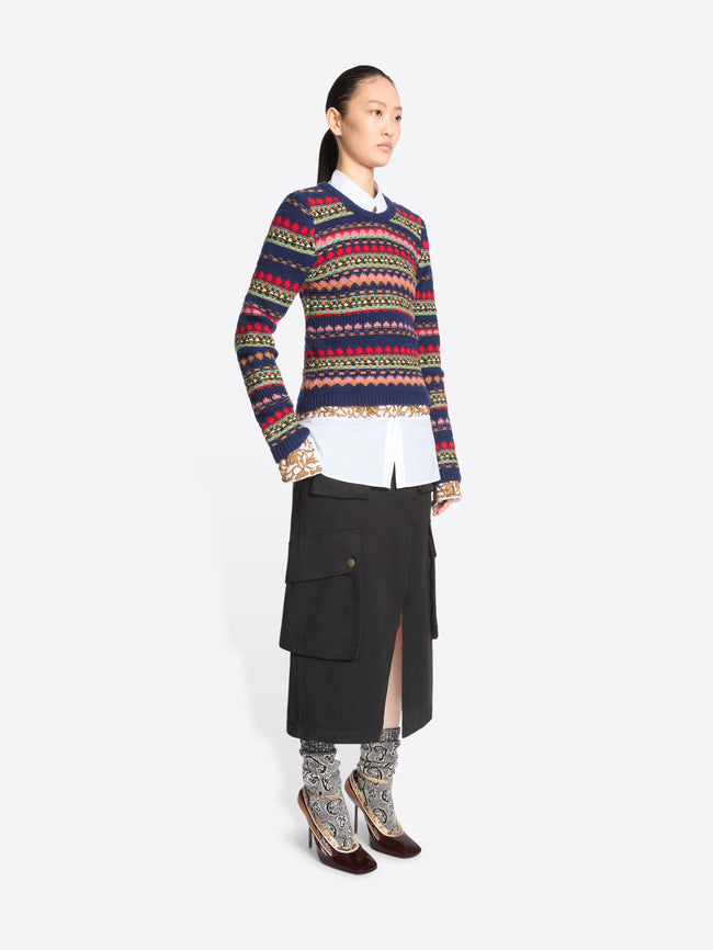 A model stands against a light gray background wearing a long-sleeve crewneck sweater with multicolor geometric stripes, a white collared shirt underneath, a black midi skirt with cargo pockets, patterned socks, and dark brown heels.