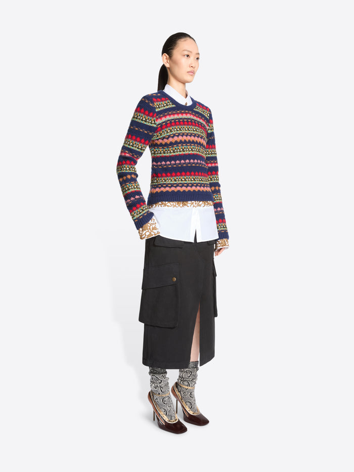 A model stands against a light gray background wearing a long-sleeve crewneck sweater with multicolor geometric stripes, a white collared shirt underneath, a black midi skirt with cargo pockets, patterned socks, and dark brown heels.