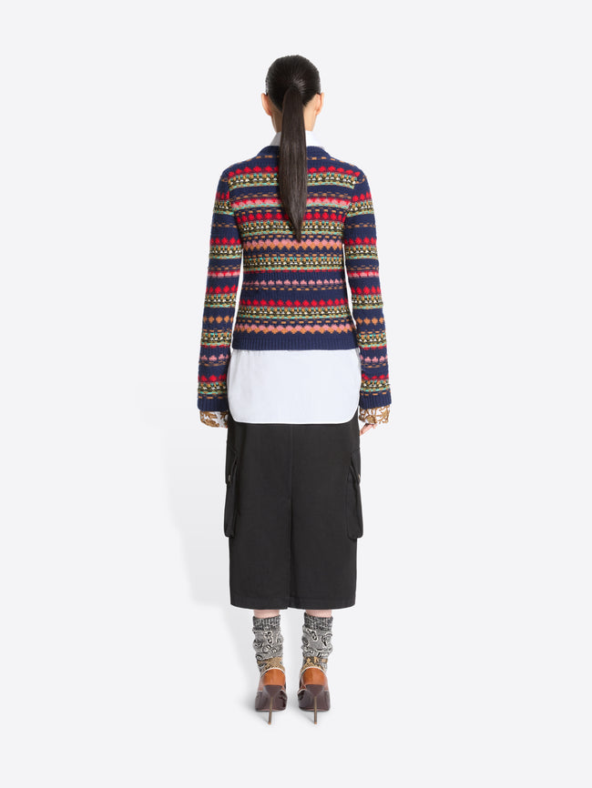 Back view of a model standing against a light gray background wearing a long-sleeve crewneck sweater with multicolor geometric stripes, a white collared shirt underneath, a black midi skirt with cargo pockets, patterned socks, and dark brown heels.