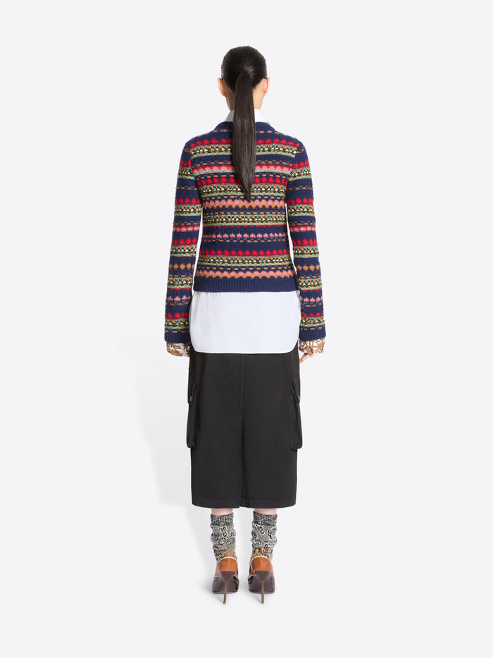Back view of a model standing against a light gray background wearing a long-sleeve crewneck sweater with multicolor geometric stripes, a white collared shirt underneath, a black midi skirt with cargo pockets, patterned socks, and dark brown heels.