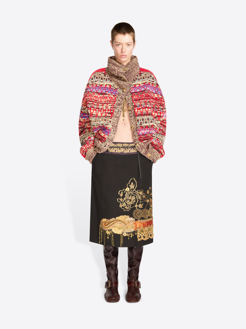 A model stands against a light gray background wearing a vibrant multicolor knit cardigan with a high collar, a black midi skirt with gold embroidery, and dark brown boots.