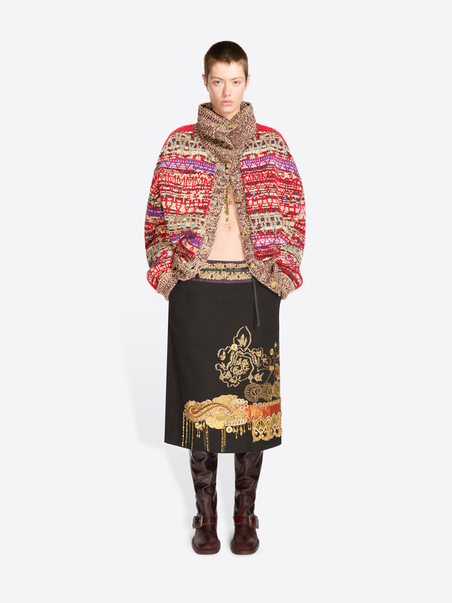 A model stands against a light gray background wearing a vibrant multicolor knit cardigan with a high collar, a black midi skirt with gold embroidery, and dark brown boots.