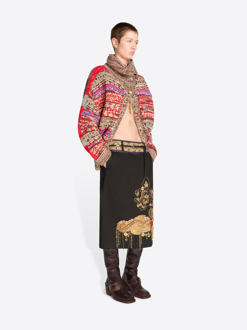 A model stands against a light gray background wearing a vibrant multicolor knit cardigan with a high collar, a black midi skirt with gold embroidery, and dark brown boots.