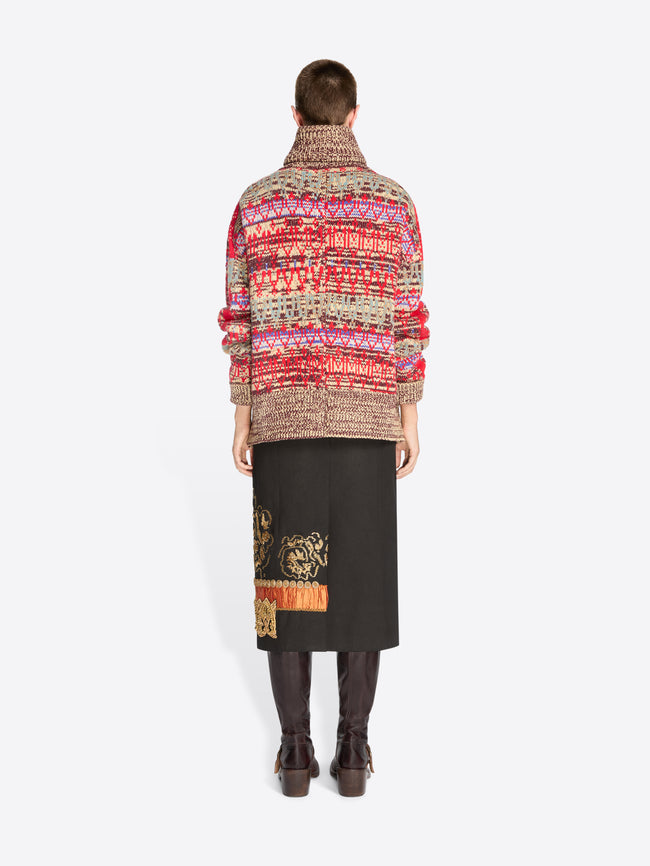 Back view of a model standing against a light gray studio background wearing a vibrant multicolor knit cardigan with a high collar, a black midi skirt with gold embroidery, and dark brown boots.