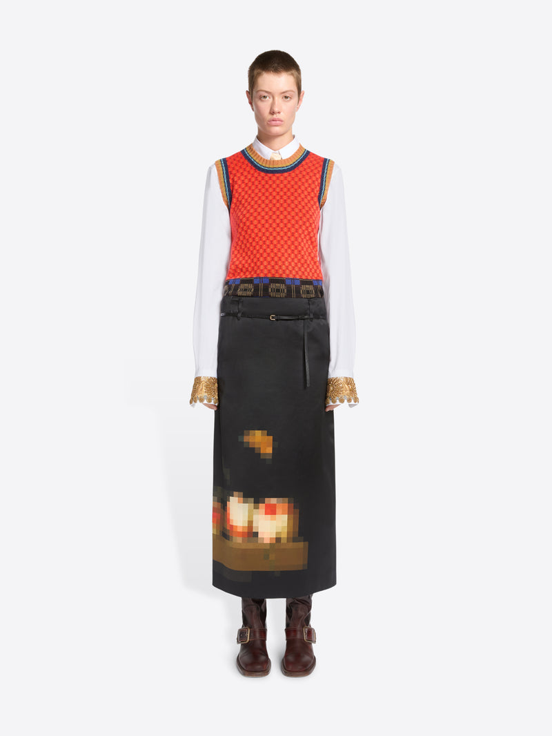 A model stands against a light gray background wearing a long black skirt with a pixelated multicolor graphic near the hem, a red patterned sleeveless top, a white long-sleeve shirt, and brown ankle boots.